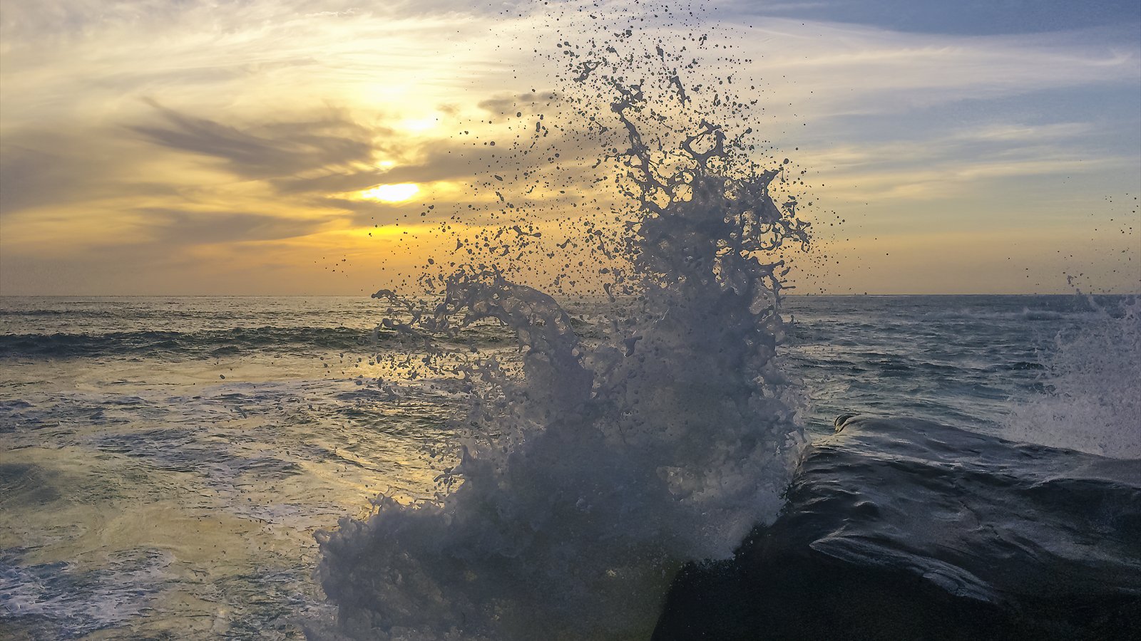 A white wave bursts upward against dark rocks at Windansea Beach as the sun sets over the Pacific.