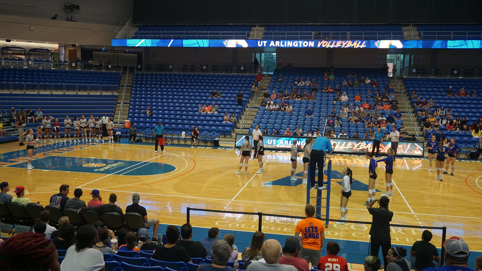 Women's college volleyball players jump at the net as teammates prepare behind them.
