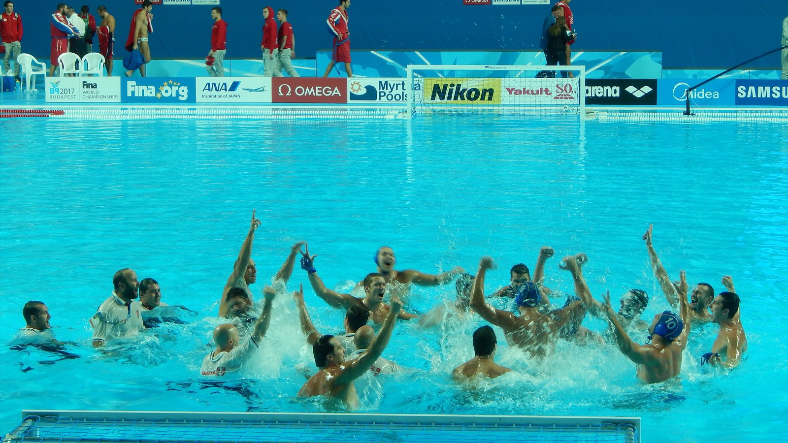 Men's water polo players and staff raise their arms while celebrating together in an indoor pool.