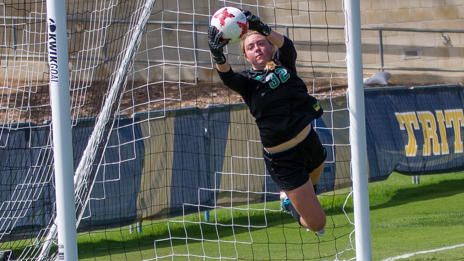 A goalkeeper catches the ball above her head at the goal line during a women's soccer match.