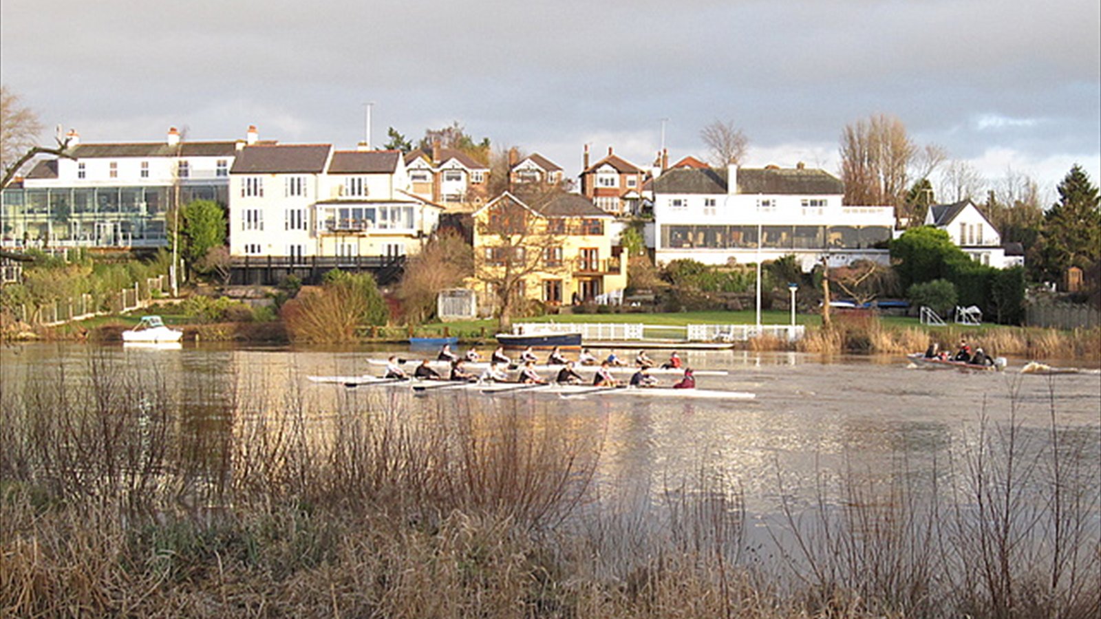 Two rowing eights race side by side on a broad river while spectators watch from the bank.