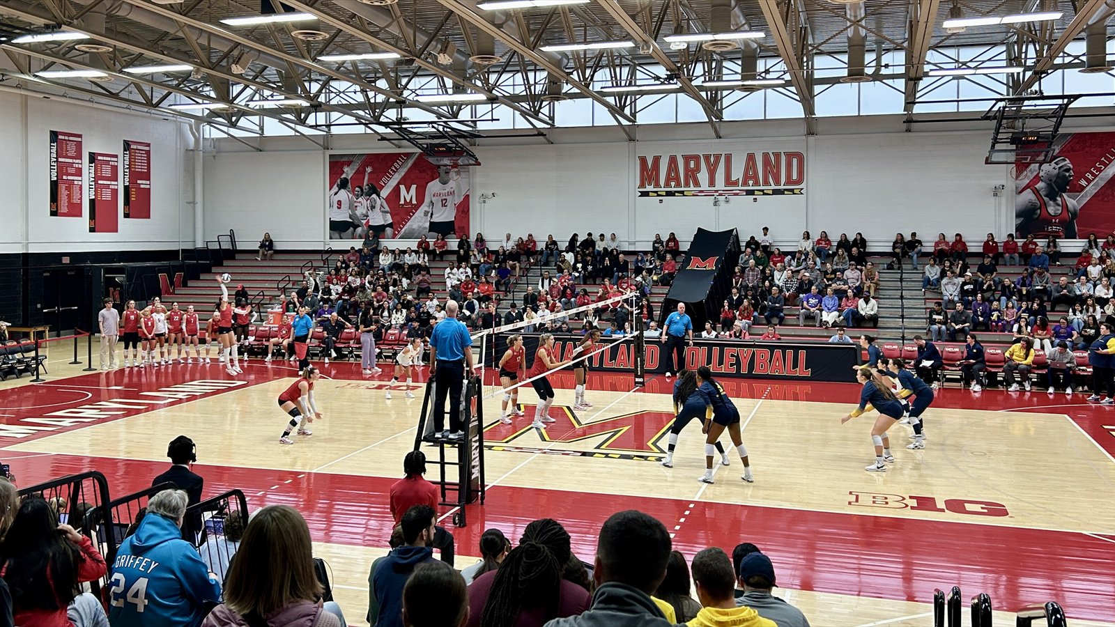 Two college women's volleyball teams face each other across the net during an indoor match.