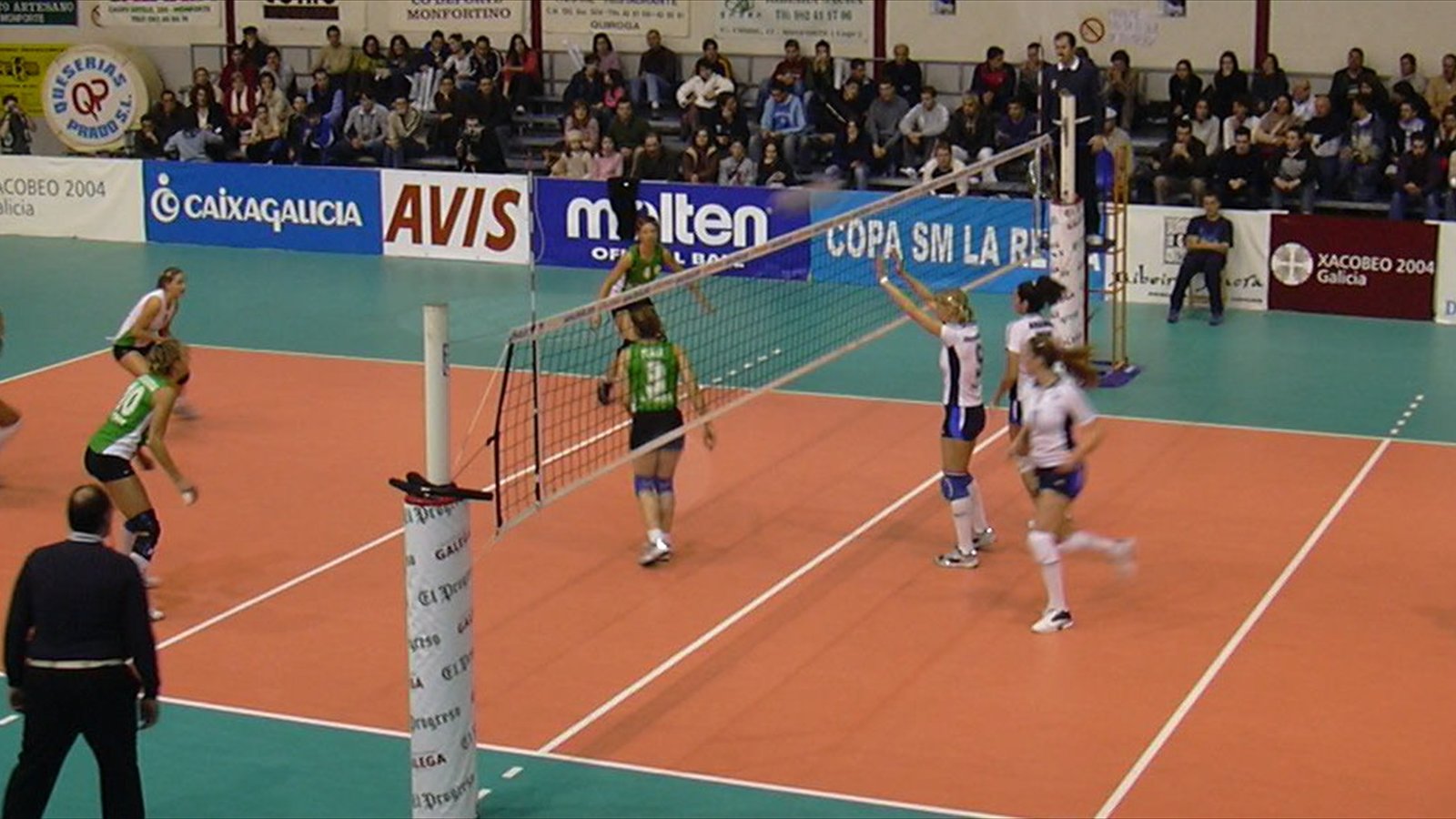 Two women's volleyball teams contest the ball above the net during an indoor match.
