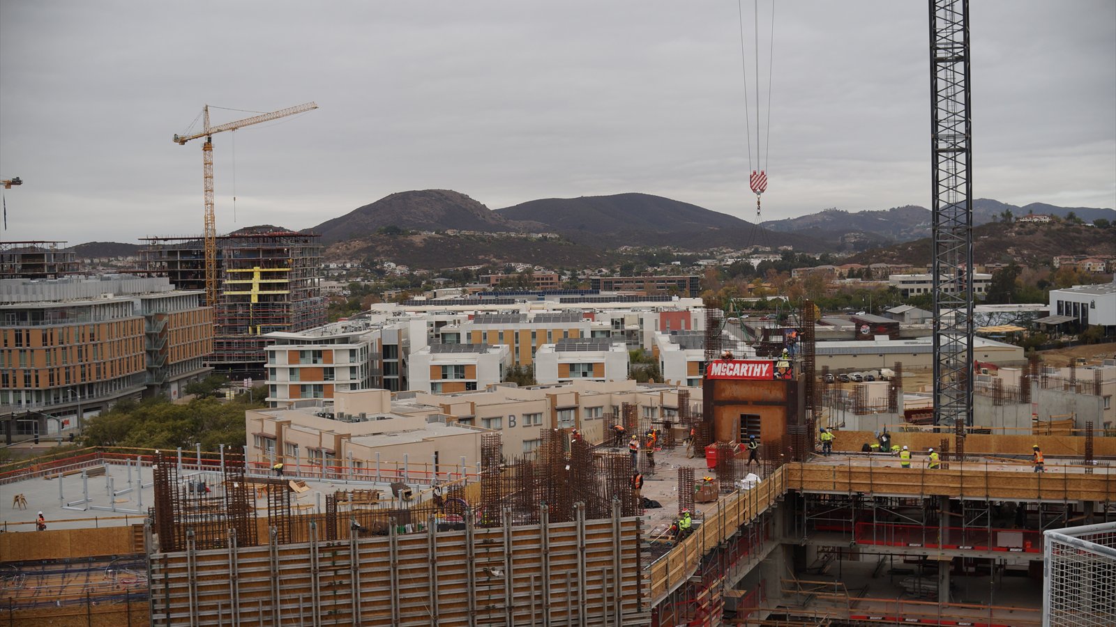 Cranes and workers surround multistory housing under construction near Cal State San Marcos, with hills in the distance.